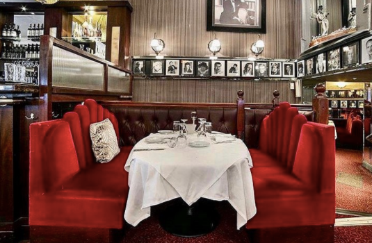 interior of trocadero restaurant. Table covered with white tablecloth, red velvet seats on either side. Black and white pictures of celebrities hanging on wall in background