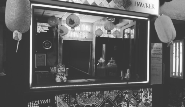 Black and white image of Hawker, a takeaway hatch with chinese lanterns, jars of food on the counter and a neon "Hang Dai" sign in the background