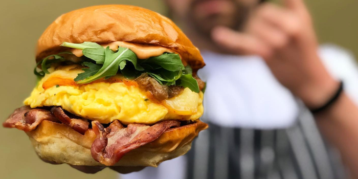 close up of a breakfast bap with rocket, soft scrambled eggs, creamy sauce and bacon. Out of focus is a man in an apron holding the sandwich