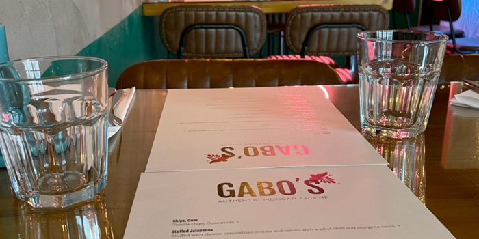 a restaurant table with glasses and a menu with the word Gabos written on top. More tables and chairs can be seen in the background