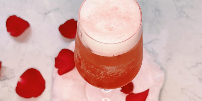 pink cocktail in a champagne glass with foam on top, with rose petals scattered in the background