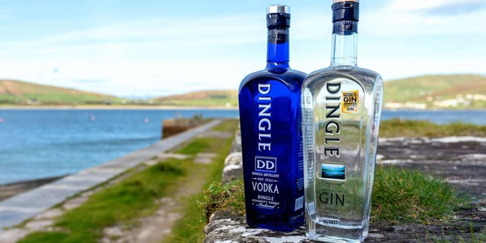 outdoor shot of a bottle of Dingle gin and Dingle vodka side by side on a grassy pier, the sea and mountains can be seen in the background