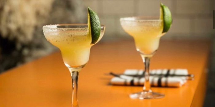 two margaritas with salt rims and fresh lime on an orange table with knives, forks and napkins beside them