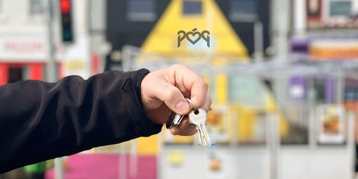 hand holding a set of keys in front of a row of shops with outdoor seating - the Póg logo can be seen in the background
