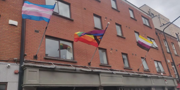 Trans flag and LGBTQ pride flag waving outside a shop front with red brick building above