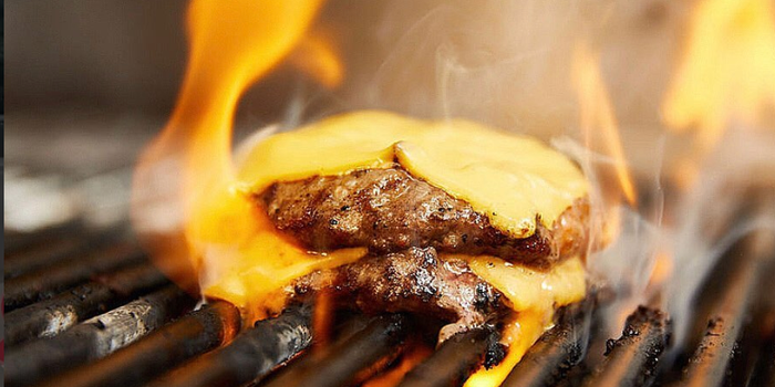 two burger patties on a flaming grill with melted cheese slices on top