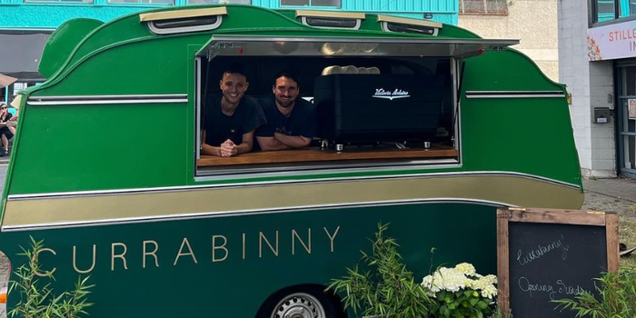 green and gold food caravan with "Currabinny" written on the side, two men standing at the hatch smiling for the camera